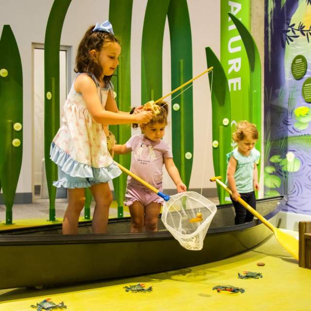 three children play in a boat and pretend to fish at the children's museum