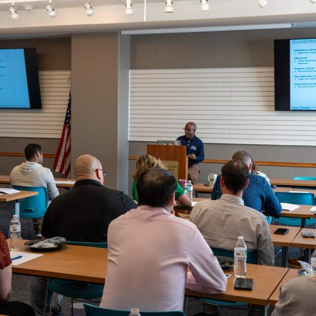 man stands in front of group in small meeting space with two screens