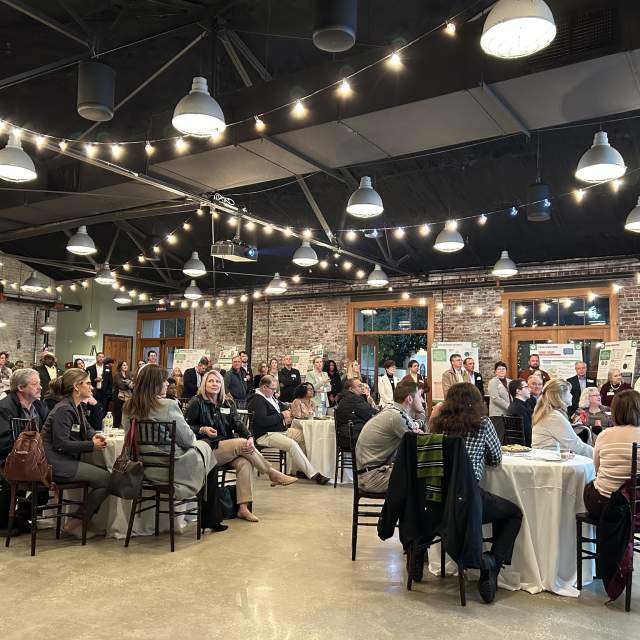large group of people sit at circle tables in an event space, twinkly lights hung from the cieling