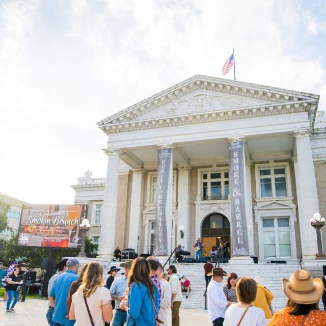 groups of people are gathered on the front lawn of the Lake Charles Courthouse. Banners hang from the columns reading smoke and barrel