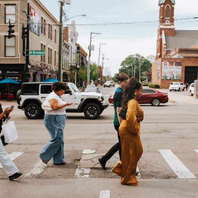 People walking across a crosswalk on a sunny day in downtown Lexington, with historic brick buildings, storefronts, and city streetlights in the background.