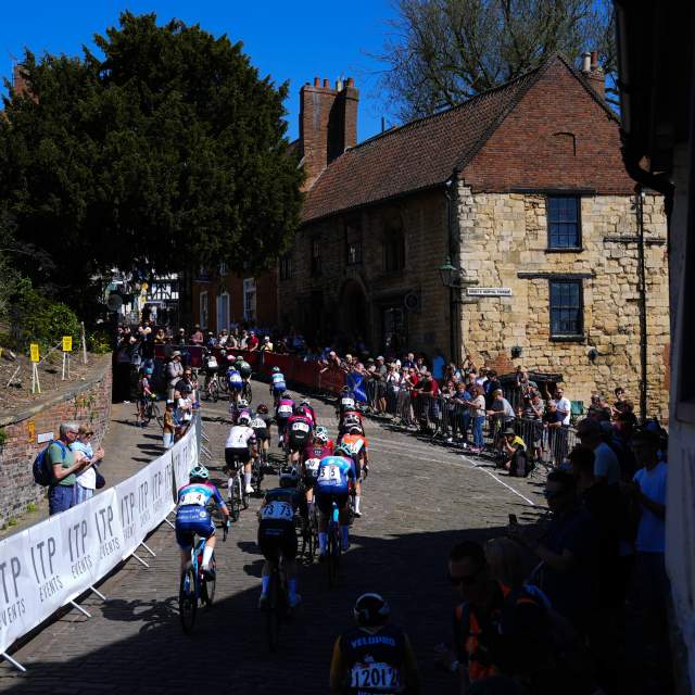 A group of cyclists compete in the Men's Tour of Britain race in Lincolnshire, with spectators watching from behind crowd barriers on either side of the route.