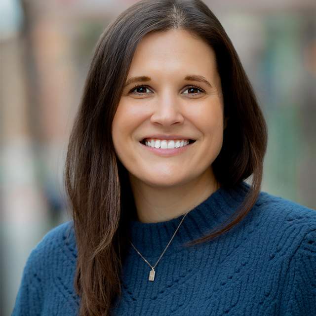 A woman with brown hair and wearing a blue sweater is smiling.