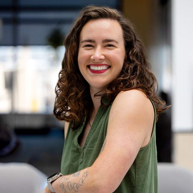 Person with shoulder-length curly hair wearing a sleeveless green top, leaning on the back of a metal chair in a bright office setting.