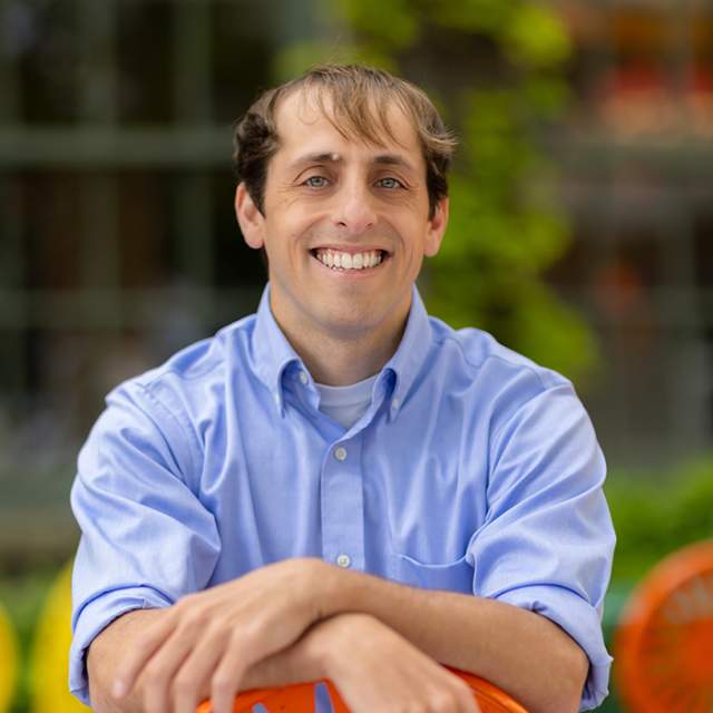 A white man wearing a blue button down shirt sitting on an orange terrace chair.