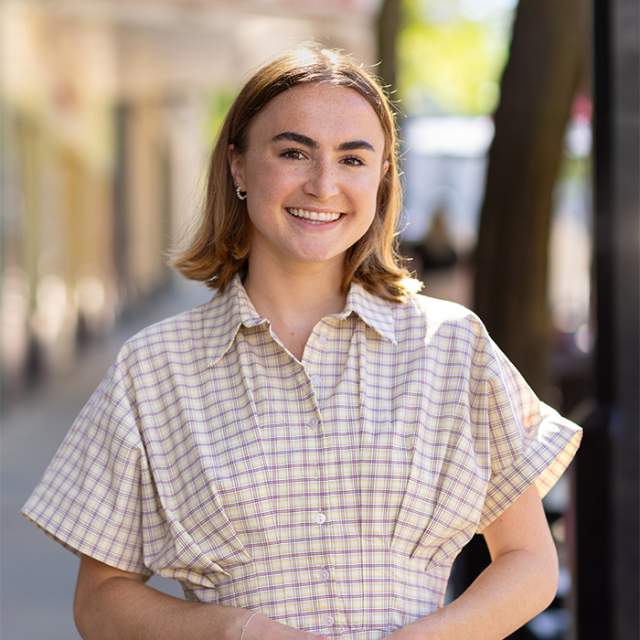 Young woman wearing a light plaid button-up dress, standing on a sunlit sidewalk with a softly blurred downtown street in the background.