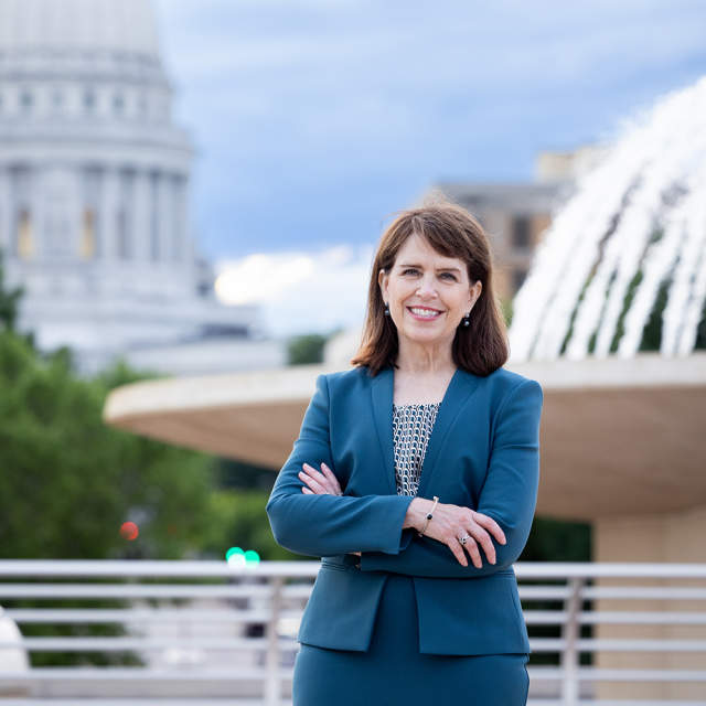 A woman with brown hair wearing a navy blue jacket, standing in front of a fountain and the Capitol building