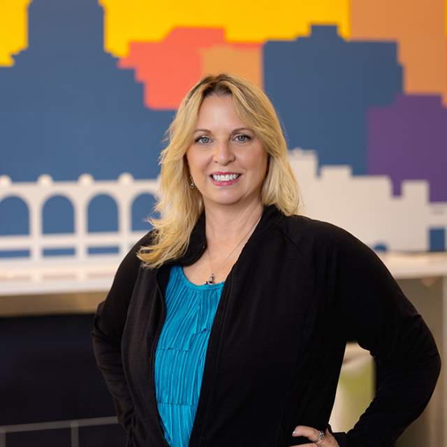 Person in a turquoise blouse and black cardigan standing with their hands on hips in front of a colorful Madison skyline mural.