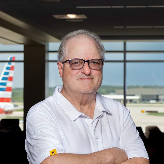 Person wearing glasses and a white polo shirt standing in an airport terminal, with an airplane visible through the windows in the background.