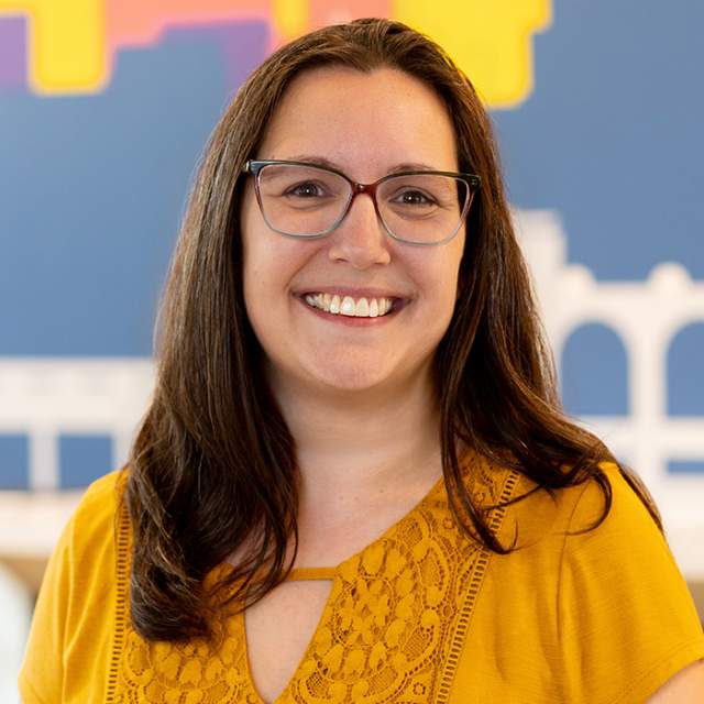Person with long dark hair and glasses wearing a mustard-yellow top, posed against a colorful blurred backdrop.
