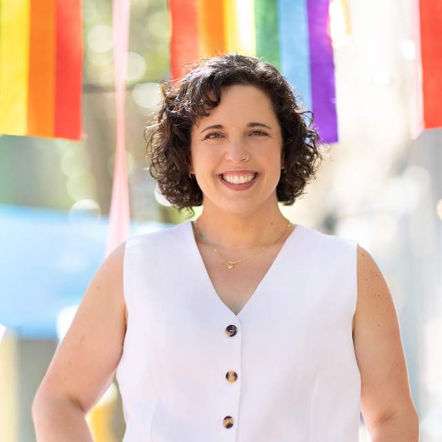 Woman wearing a white sleeveless button-front top, standing outdoors beneath colorful rainbow ribbons with a softly blurred background.