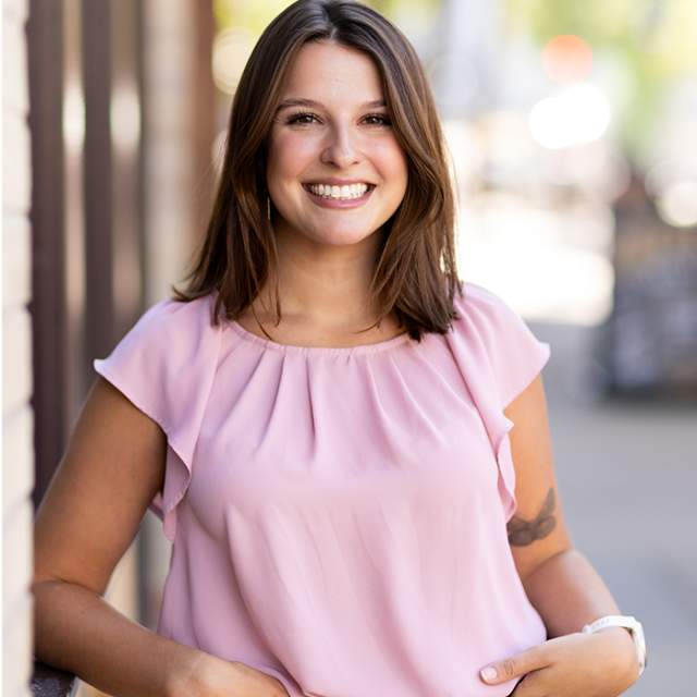 Young woman wearing a light pink blouse and dark pants, standing on a downtown sidewalk with a softly blurred street background.