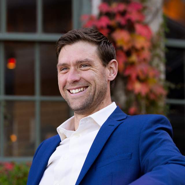 A white man wearing a white shirt and blue blazer sitting down and smiling in front of a window with foliage.