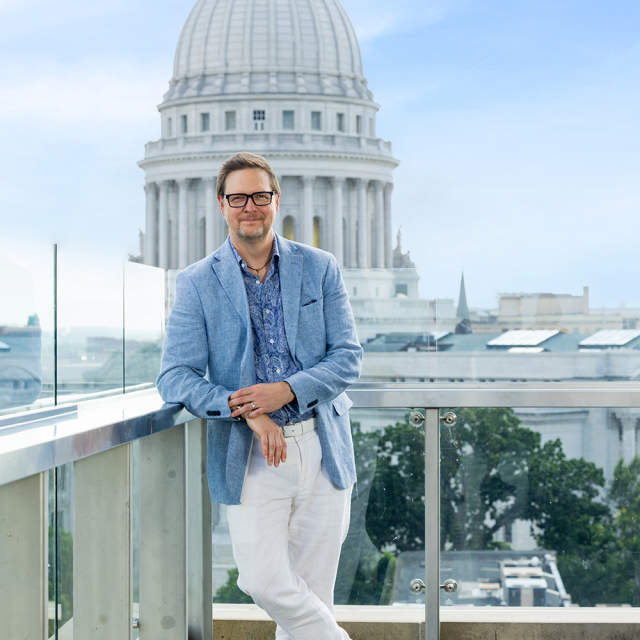 A man wearing a blue button up, blue suit jacket and white pants standing on a rooftop balcony with the state capitol building behind him.
