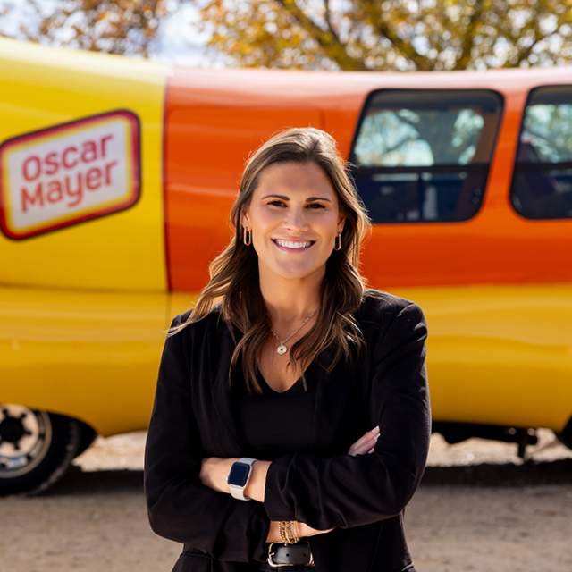 A white woman with light brown hair is wearing a black shirt underneath a black blazer with her arms crossed and smiling while posing in front of the Oscar Mayer Weinermobile.