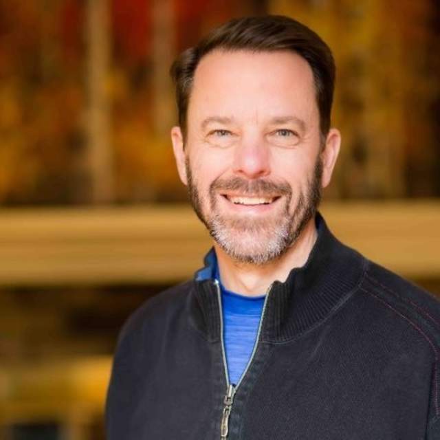 A white man with brown hair wearing a navy blue jacket smiling.
