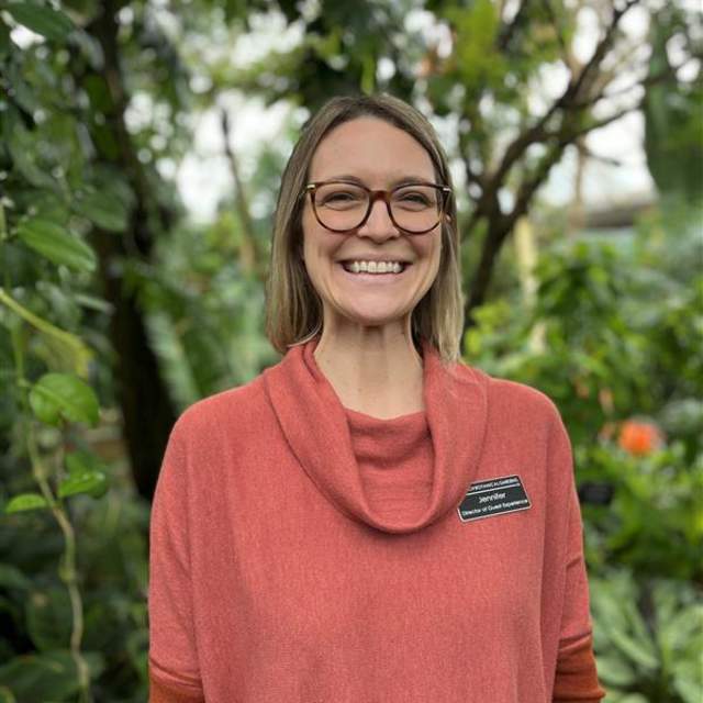 A woman with short hair and glasses standing in front of green trees.