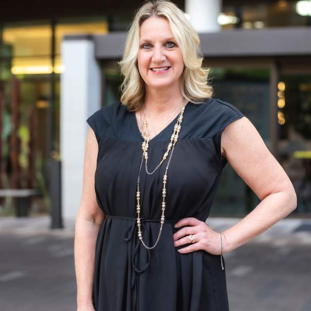 A white woman with blonde hair wearing a black dress stands in front of building downtown.