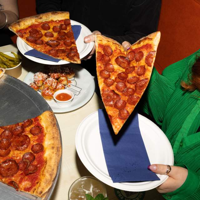 A large pepperoni pizza is being served at a restaurant, with several slices being handed out on white plates. A woman in a green top holds a plate as a slice is lifted from the pizza, while dipping sauces and drinks sit on the table nearby.