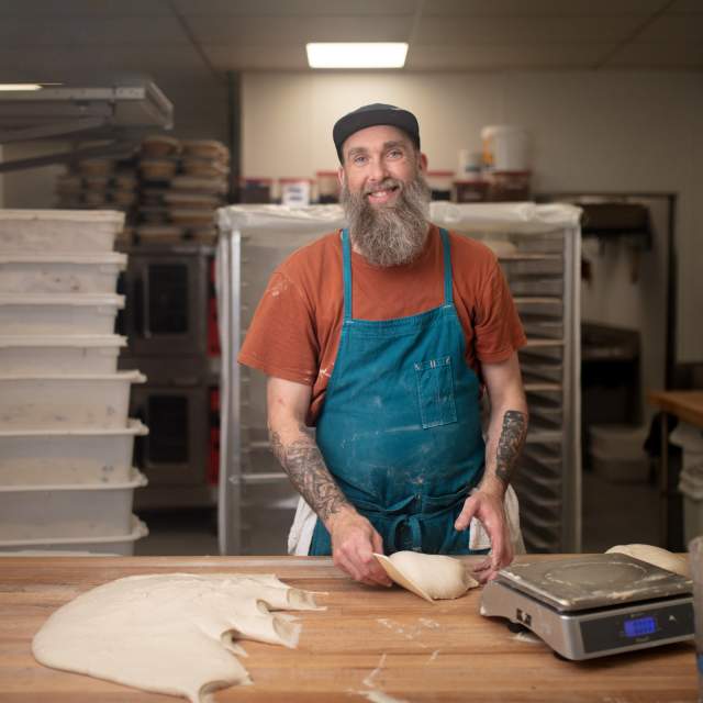 Photo of a bearded man in a rust-colored tee shirt and blue apron, working with bread dough on a big cutting board