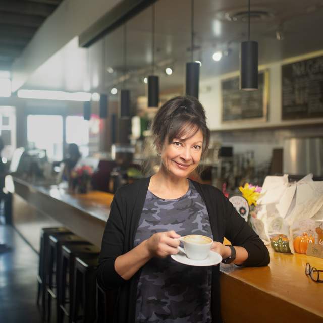 A smiling brunette woman standing at a counter holding a coffee cup