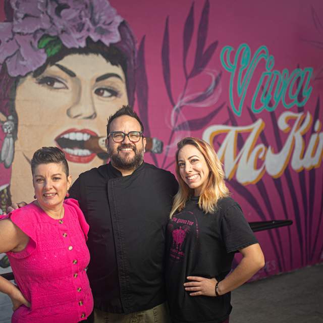 two women and man standing in front of a pink mural of a woman that says Viva McKinney