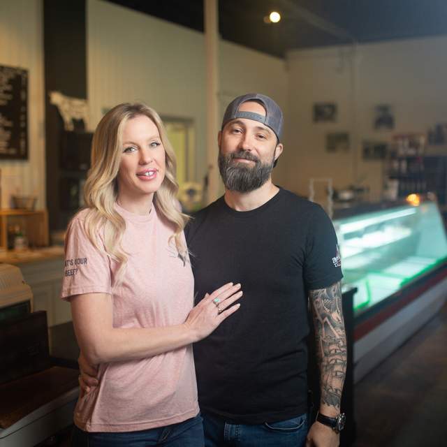 blonde woman in pink shirt with bearded man in black tshirt and tattooed arms in from of meat case
