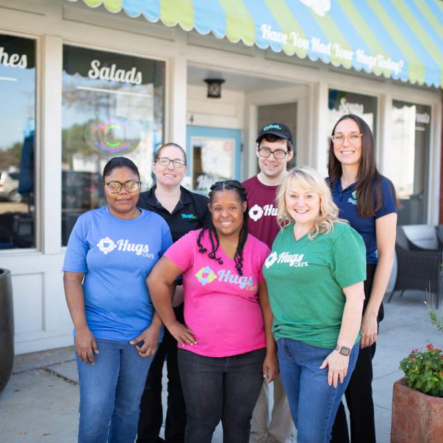 Diverse group of people wearing various colored Hugs tshirts in front of cafe