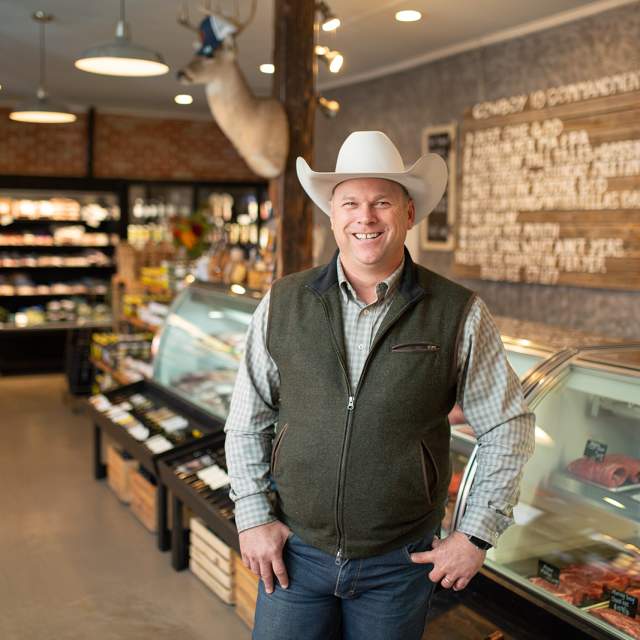 Man in a cowboy hat, long sleeves, and a zippered vest standing in front of a meat counter full of cuts of beef