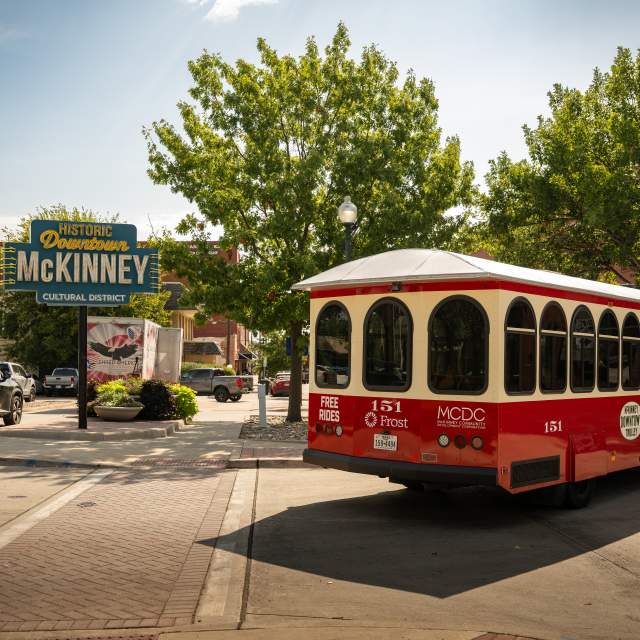 Red and white Downtown McKinney trolley driving past the neon Historic Downtown McKinney sign.