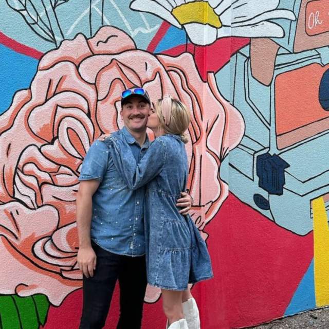 A young blonde woman in a denim dress and white boots kisses a young mustached man on the cheek in front of a colorful mural