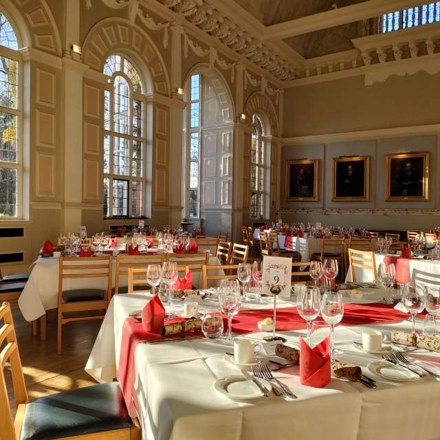 Newnham College Cambridge College dining hall set for a Christmas banquet. The walls are light & there is autumnal light streaming through the windows. The tables are fully set up with white linen, scarlet table runner, silverware and glasses