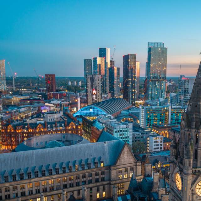 A panoramic evening view of Manchester city centre at dusk, with the illuminated clock tower of Manchester Town Hall Extension in the foreground and Manchester Central Convention Complex at the heart of the skyline. Modern high-rise towers at Deansgate Square and surrounding city lights glow against a blue twilight sky, showcasing the contrast between Manchester's historic and contemporary architecture.
