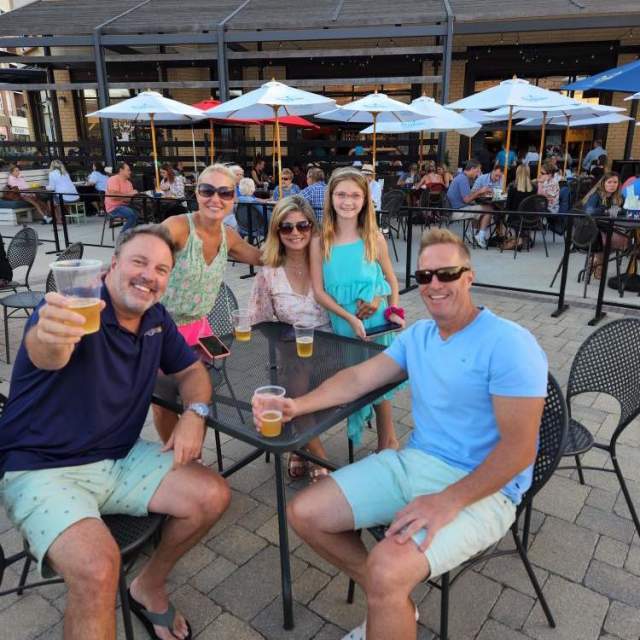 A group of five people, including two men, two women, and one girl, smile and pose for a photo while sitting at an outdoor patio table with drinks. They are surrounded by other diners under white and blue umbrellas, enjoying a sunny day. One man raises his drink in a cheerful toast.