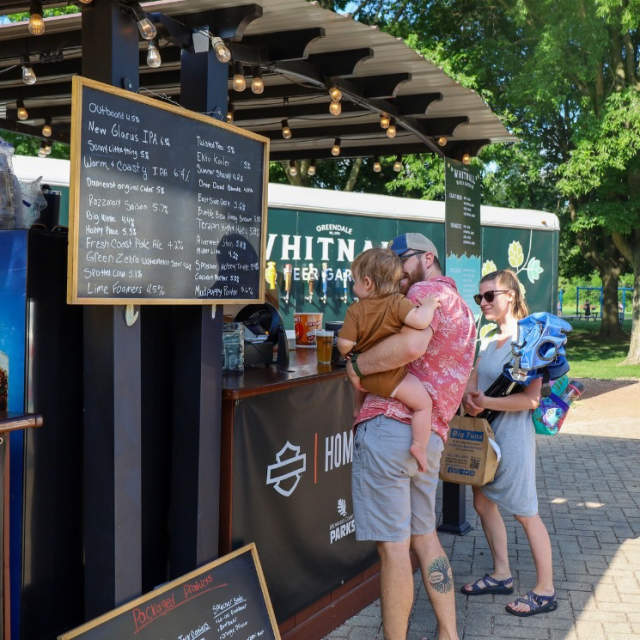 A father holding his child next to his partner at a beer garden counter ready to order some food or drinks in a summer setting with trees in the background.