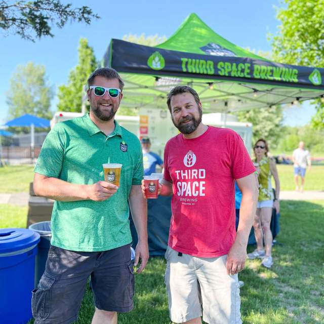 Two men, on the left wearing a green shirt holding a beer, on the right wearing a red shirt holding a beer, in front of a tent with a navy roof in a park with green trees and grass in the background.