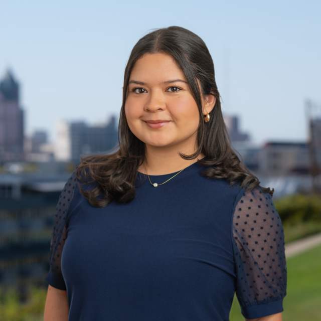 Chelsea Hernandez smiling at the camera with the Milwaukee skyline softly blurred in the background. She poses on a grassy overlook under a clear blue sky.