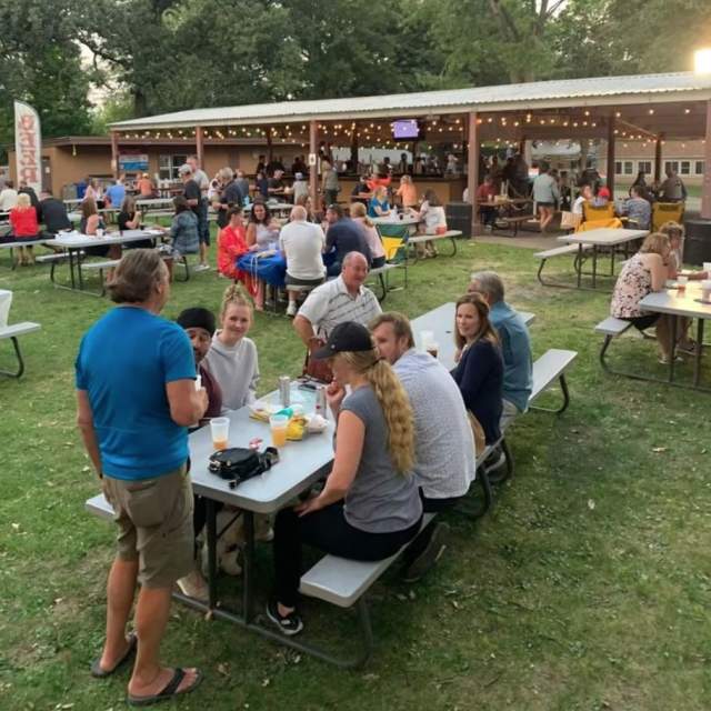 A group of people sitting at a picnic table enjoying a meal and some beer at dusk in a beer garden setting.
