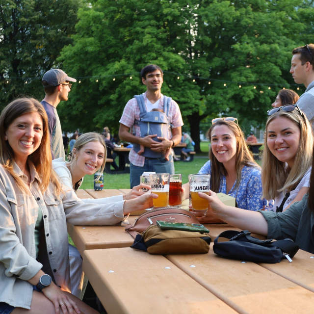 A group of friends sitting at a park picnic table smiling at the camera as they raise their glasses to cheers, with a group of men talking behind them, with trees in the background.