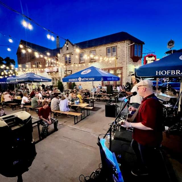 A wide, nighttime view of the Kegel's Inn patio, with string lights illuminating the space, as musicians play their instruments in the foreground and diners enjoy food and drinks in the background.