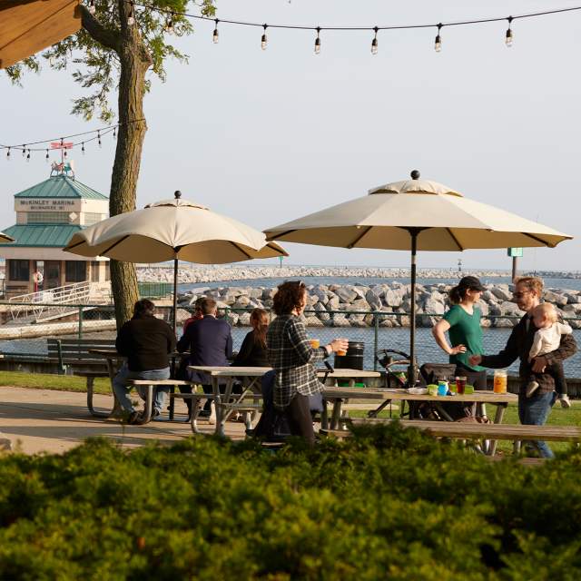 A group of people enjoying beers under umbrellas at the picnic tables outside of the Roundhouse Beer Garden in Milwaukee along the Lakefront.