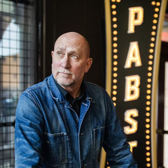 Gary Witt sits wearing a denim shirt in front of an illuminated Pabst sign with vintage-style light bulbs, with large windows visible in the background