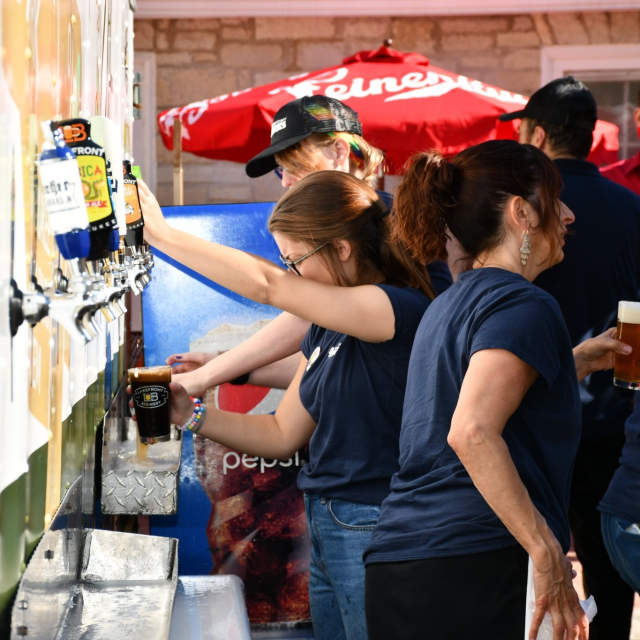 A group of beer garden employees behind the counter pouring beers from tap handles on a sunny day.