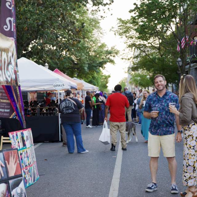 An eye-level, slightly wide shot captures a lively street market during the late afternoon. In the foreground, a man  stands holding a plastic cup, chatting with a woman. To their left, a henna tattoo booth is decorated with posters, with an artist seated inside. Beyond them, a row of white vendor tents lines the street, with a crowd of people strolling through the market.