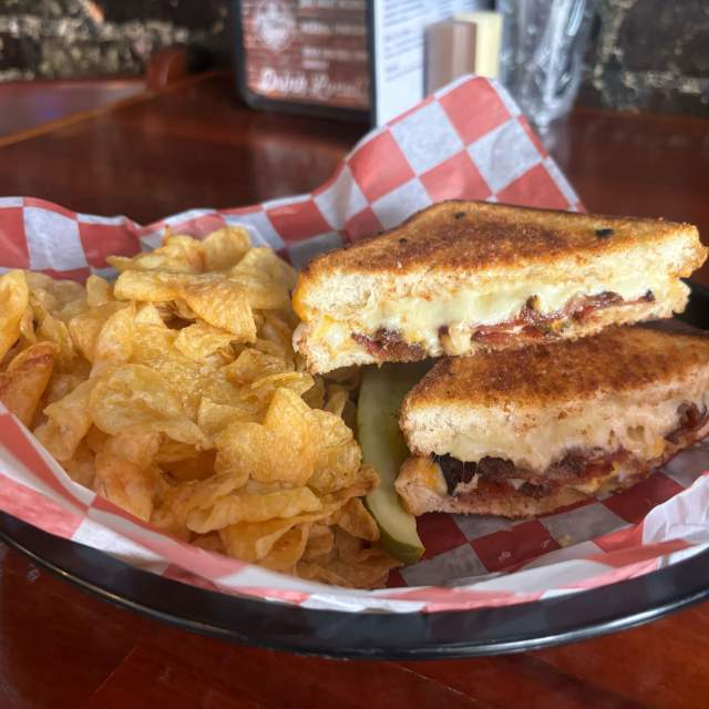 Grilled cheese with bacon, cut in half with chips and a pickle in a basket with red and white checkerboard paper