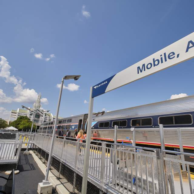 Amtrak train at the Mobile, Alabama station with passengers boarding, set against the downtown skyline including the RSA Tower and Arthur R. Outlaw Convention Center.