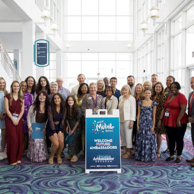 About two dozen people pose for a group photo behind a sign that says Visit Mobile Welcome Tourism Ambassadors