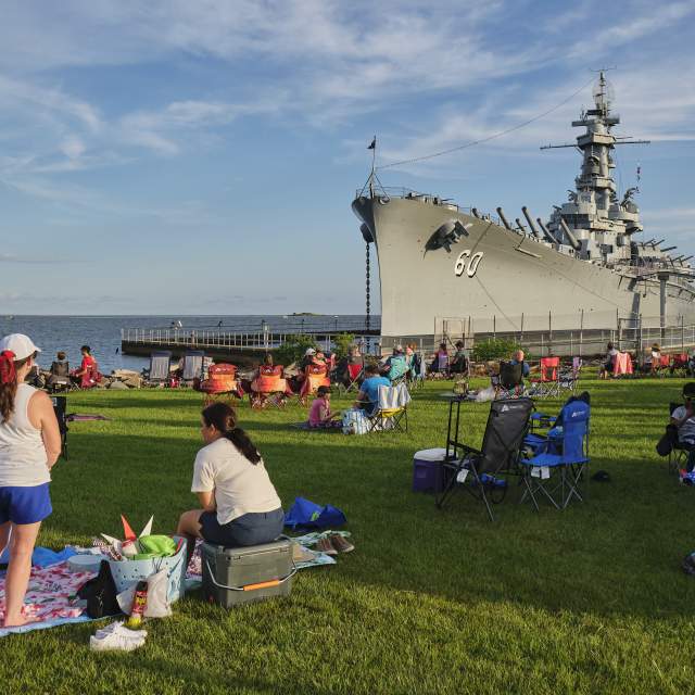Crowd enjoying a sunny day on the grass near a large battleship, with a ramp leading to the ship and an American flag backdrop.