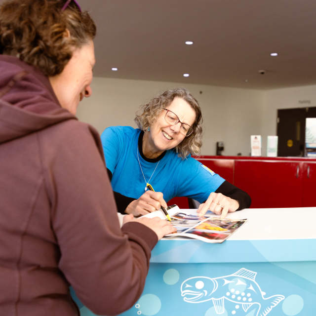 A visitor receives help from staff at a tourism information desk while reviewing a brochure together.