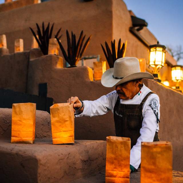 fernando olea, in chef attire and a cowboy hat adjusting a luminario or traditional paper bag with a flame inside